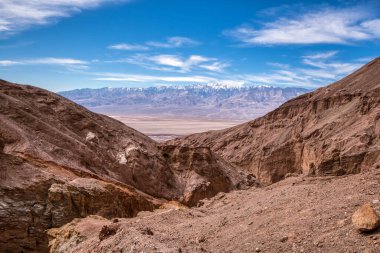 Engebeli kanyon duvarları uzak karlı tepeleri çerçeveliyor. İnce bulutlu mavi gökyüzü dingin bir kontrastı ekler. Kuru, desenli toprak, kurak ve el değmemiş vahşi doğayı işaret ediyor. Geniş vadi aşağıda uzanır.