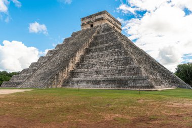 Ancient pyramid rises under bright blue sky. Stone steps lead to temple atop sacred ground. Clouds drift gently over historical wonder. Grass blankets base of massive monument