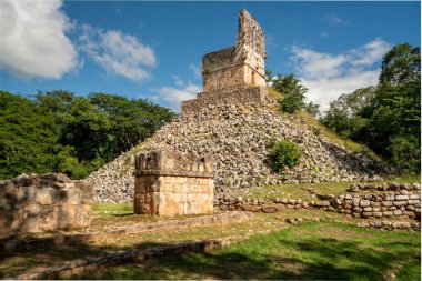 Ancient stone pyramid rises from green jungle floor. Sky bright blue with scattered clouds above. Lush trees surround the sacred ruins. Sunlight casts warm glow on weathered stones