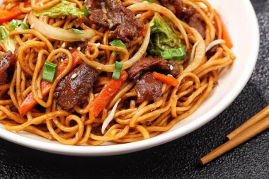 Bir tabakta sığır eti, kızarmış erişte, taze et dilimleri ve baharatlı soslu karışık sebzeler servis ediliyor.