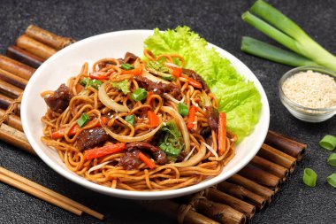 Bir tabakta sığır eti, kızarmış erişte, taze et dilimleri ve baharatlı soslu karışık sebzeler servis ediliyor.