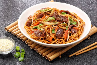 Bir tabakta sığır eti, kızarmış erişte, taze et dilimleri ve baharatlı soslu karışık sebzeler servis ediliyor.