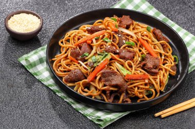 Bir tabakta sığır eti, kızarmış erişte, taze et dilimleri ve baharatlı soslu karışık sebzeler servis ediliyor.