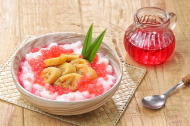 Bir kase renkli tatlı, muz, kırmızı şurup ve ahşap masa üzerinde yeşil yapraklar.