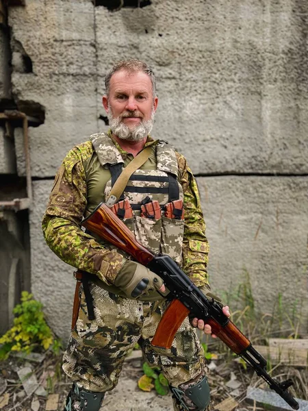 Kamuflaj üniformalı orta yaşlı asker saldırı tüfeğini nişan alıyor, savaş alanında taktik eğitimi alıyor..