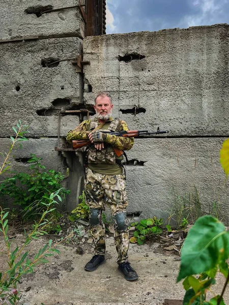 Kamuflaj üniformalı orta yaşlı asker saldırı tüfeğini nişan alıyor, savaş alanında taktik eğitimi alıyor..