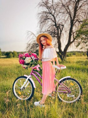 Büyük bir buket şakayıkla süslenmiş eski bir bisikletle hasır şapkalı güzel bir genç kız. Gün batımında altın saatle büyüleyici kırsal manzara.