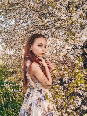 Çiçekli taç takmış güzel kızıl bir kızın yakın plan portresi. Yüzüne dokunuyor ve kameraya bakıyor. Çiçek açan bir bahçede yumuşak altın saat aydınlatması.