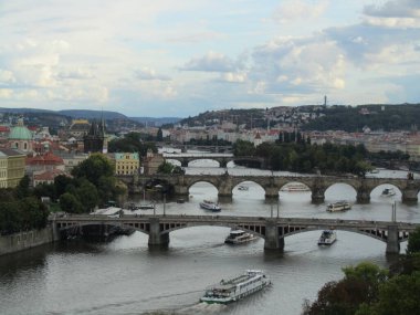 Vltava Nehri 'ni kaplayan ikonik Charles Köprüsü' nün birden fazla köprüsü ve tarihi Prag şehri manzarası. Bulutlu gökyüzünün altında seyreden turist tekneleri.