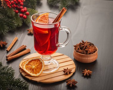 Ahşap bir masada birkaç portakal dilimi ve tarçınlı sıcak şarap. Yakınlarda kırmızı böğürtlen ve anason yıldızlı ladin dalları var..