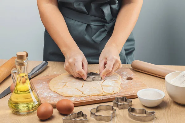 A girl in a gray apron without a face cuts hearts out of shortbread dough with an iron mold. On the table are a wooden rolling pin, flour, oil in a glass bottle, a knife, eggs, sugar and parchment. Lifestyle.