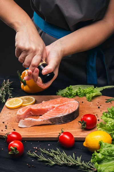 Cooking salmon. Female hands sprinkle salmon fish steak with spices on a beautiful wooden board. Ingredients lemon, rosemary, peppercorns, tomatoes, peppers, lettuce. Close-up, side view. Wellness