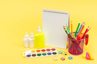 Bardaktaki renkli kalemler, bir defter, yapıştırıcı, zımba, boya, renkli ataçlar ve sarı arkaplanda bir silgi. Kopyalayacak bir yer. Reklam ve tasarım için bir boşluk. Mükemmel rötuş..