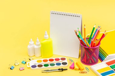 Bardaktaki kalemler, yaydaki bir defter, yapıştırıcı tüpleri, zımba, boyalar, plastikler, defterler, pusulalar, ataçlar ve sarı arkaplanda bir silgi. Boşluğu kopyala.