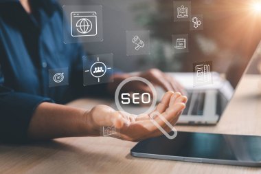 Modern çalışma alanında bilgisayar ve tablet üzerinde SEO hologram simgelerini açık elle sunarak dijital pazarlama stratejisini, web sitesinin optimizasyonunu, analitiğini, anahtar kelime araştırmasını ve çevrimiçi büyümeyi sembolize ediyor.