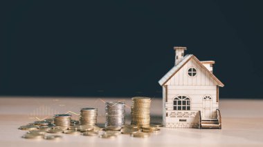 Yığılmış madeni paraların yanında ahşap ev modeli, masanın üzerindeki ince piyasa çizelgesi, emlak yatırımlarını, ev tasarruflarını, ipotek planlarını, finans stratejisinde yükselen gayrimenkul değerini gösteren çizimler.