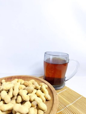 Bambu hasırının üzerindeki tahta bir tabak fıstığın yanında ılık bir bardak bitki çayı, beyaz bir arka plana kurulmuş, basitliği, sağlığı ve geleneksel ferahlığı sembolize ediyor.