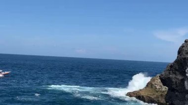 Sakin bir mavi denizde yatay hareket eden küçük bir tekne kayalık bir kıyı şeridinden çekilerek huzurlu bir sahil atmosferi yakalıyor.