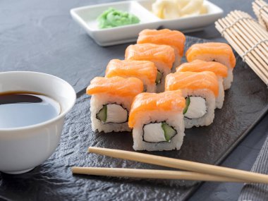 Suşi Philadelphia 'yı gri arka planda siyah desenli bir tabakta yuvarlıyor. Wasabi zencefili ve sos çubukları. Somon, krem peynir, avokado..