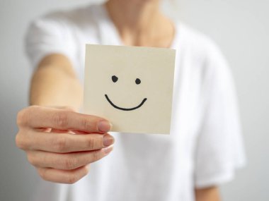 Genç bir kadın, uzanmış elinde çizilmiş gülen yüzlü kağıt bir çıkartma tutuyor. El ve yapıştırmaya odaklan, bulanık arkaplan