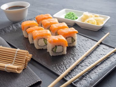 Suşi Philadelphia 'yı gri arka planda siyah desenli bir tabakta yuvarlıyor. Wasabi zencefili ve sos çubukları. Somon, krem peynir, avokado..