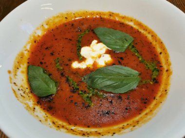 Fesleğen yapraklarıyla süslenmiş beyaz bir tabakta Stracciatella peynirli İtalyan domates çorbası.