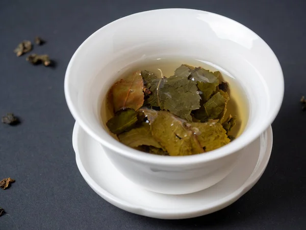 a white bowl with brewed tea leaves on a dark background. Close up, side view. Chinese tea, open leaves