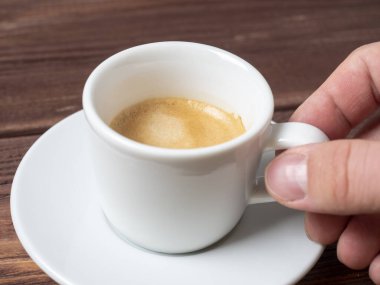 Beyaz bir çay tabağının üzerinde beyaz bir fincan aromatik espresso tutan bir adamın yakın çekimi. Ahşap zemin, kenar görünüm.