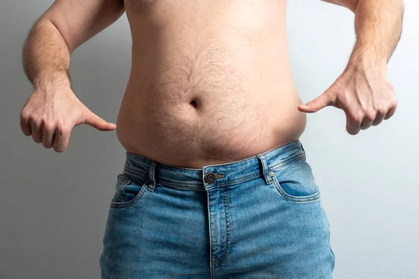 a fat man in jeans with a bare stomach shows his thumbs down with both hands.