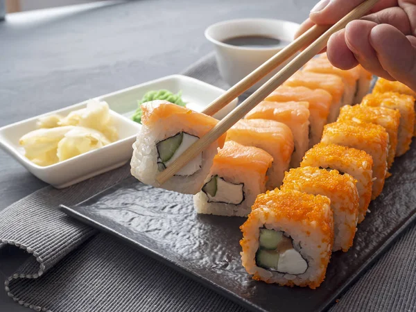 Philadelphia ve California desenli siyah bir tabakta yuvarlanıyor. Bir rulo, diğerlerinin üstündeki büyükanne çubuklarında tutuluyor. Soya sosu, wasabi ve zencefil. Yan görüş. Japon mutfağı.