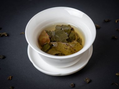 Siyah arka planda demlenmiş çay yaprakları olan beyaz bir kase. Yaklaş, yan görüş. Çin çayı, açık yapraklar.