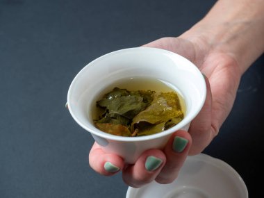 Bir kadının eli beyaz bir porselen kase ile koyu arkaplanda demlenmiş yeşil çay tutar.