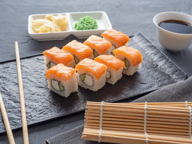 Suşi Philadelphia 'yı gri arka planda siyah desenli bir tabakta yuvarlıyor. Wasabi zencefili ve sos çubukları. Somon, krem peynir, avokado..