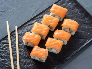 Philadelphia suşisi gri arka planda siyah desenli bir tabakta yuvarlanıyor. Yakınlarda bambu çubukları var. Üst manzara, düz yatış. Japon mutfağı.