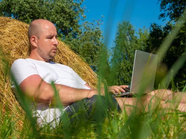a view from below through the grass of a man sitting near a haystack with a laptop. Remote work, rural area