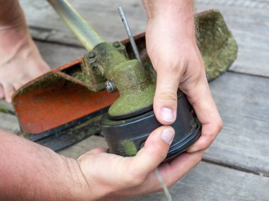 Yakından çekilen erkek eli, benzin düzenleyicisinde olta ipiyle başlığı söküyor. Bahçe malzemelerinin bakımı ve onarımı