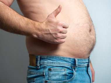 Gri arka planda kot pantolon giymiş koca göbekli bir adam. Kaldırılmış bir parmak. Kendini kabullenme, vücut pozitifliği.