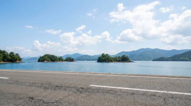 Sakin mavi sulara dağılmış küçük adalarla dolu bir sahil yolu manzarası, uzak dağlar tarafından çerçevelenmiş ve tüylü bulutlarla dolu parlak bir gökyüzü..