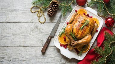 Portakal dilimleri ve kızılcıklarla süslenmiş güzel kavrulmuş bir tavuk, kırsal bir ahşap masada beyaz bir tepside oturur. Sahne, çam dalları, kırmızı süsler ve altın boncuklar da dahil olmak üzere şenlikli Noel süslemeleriyle süslenmiş.