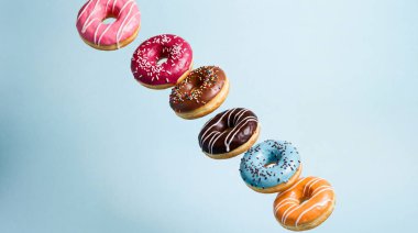 Her biri farklı renkte şekerleme ve serpiştirilmiş altı donuttan oluşan canlı bir dizi havada yakalanıyor. Çapraz olarak mavi bir zemin üzerinde diziliyor ve neşeli ve iştah açıcı bir görsel ortam yaratıyor..