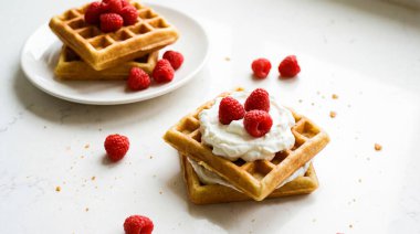 Lezzetli bir yığın altın-kahverengi waffle, cömertçe süslenmiş canlı kırmızı ahududu ve bir tutam bembeyaz krema, temiz beyaz bir yüzeyde sunuluyor. Arka planda ahududulu bir tabak daha waffle görünüyor, kreati.