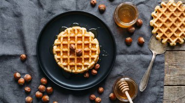 Koyu bir tabakta servis edilen, altın bal serpiştirilmiş ve fındıkla süslenmiş yeni pişmiş waffle 'ın üst görüntüsü. Bir kavanoz bal ve dağınık fındık kırsal kahvaltı sahnesini tamamlıyor..