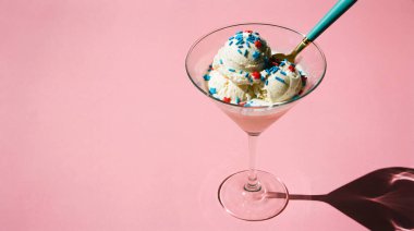 Bir kepçe vanilyalı dondurma ile dolu yüksek açılı bir martini bardağı, cömertçe üzerine mavi ve kırmızı serpiştirilmiş, ve mavi bir kaşık. Cam parlak pembe arka planda uzun bir gölge bırakıyor, neşeli ve davetkar bir tatlı yaratıyor.