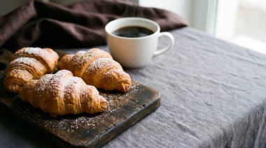 Altın esmer kruvasanların toz şekerle kaplandığı enfes bir kahvaltı sahnesi, kırsal bir ahşap kesme tahtasına ustaca yerleştirilmiş, yanında yumuşak, doğal bir ışık arka planına karşı hazırlanmış, dumanı tüten bir fincan koyu kahve ile birlikte..