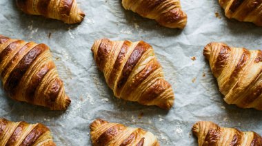 Birden fazla altın-kahverengi kruvasan resmi, mükemmel pişirilmiş ve buruşuk parşömen kağıdının üzerinde sıralanmış, ince dokularını ve zengin renklerini vurgulayan..