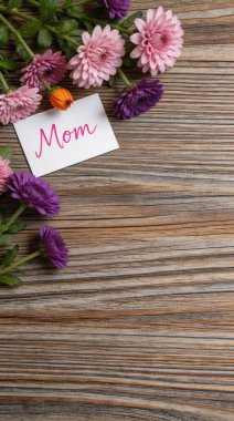 Canlı pembe ve mor çiçeklerin arasına yerleştirilmiş içten bir anne kartı. Dokulu ahşap bir yüzeye ustaca yerleştirilmiş. Anneler Günü ya da doğum günü için mükemmel..