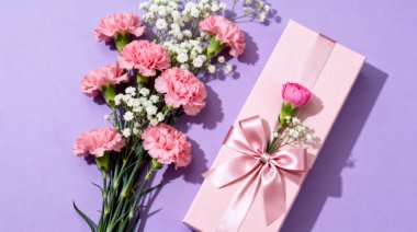 Pembe karanfiller ve narin beyaz bebek nefesli çiçekler, pembe bir hediye kutusunun yanında saten bir kurdeleyle bağlanmış, canlı mor bir yüzeye kurulmuş, kutlamalar için mükemmel..