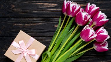 Parlak pembe lalelerin çarpıcı bir üst çekimi düzgün paketlenmiş bir hediye kutusuyla eşleştirilmiş, hepsi de kırsal, karanlık, ahşap bir yüzeye karşı, kutlamalar veya özel durumlar için mükemmel..