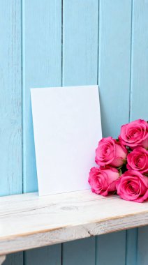 Açık ahşap bir rafta duran el değmemiş beyaz bir kart. Yanında da büyüleyici mavi ahşap bir arka plana kurulmuş canlı pembe güller var..