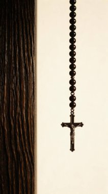 Siyah boncuklu ve gümüş haçlı bir tespihin yakın plan çekimi, dikey olarak açık renkli arka planda asılı, sol tarafında koyu ahşap bir doku var..